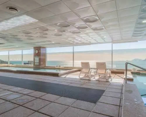 Busan Haeundae HILL SPA's hot spring area, featuring various temperature pools and lounge chairs, offering an unbeatable Haeundae sea view through large glass windows for ultimate relaxation.