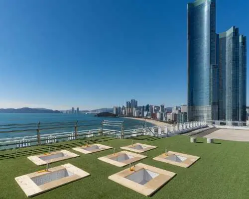 Busan Haeundae HILL SPA's rooftop outdoor relaxation area, featuring green artificial turf with square seating spaces, overlooking the vast ocean and Haeundae city skyline.