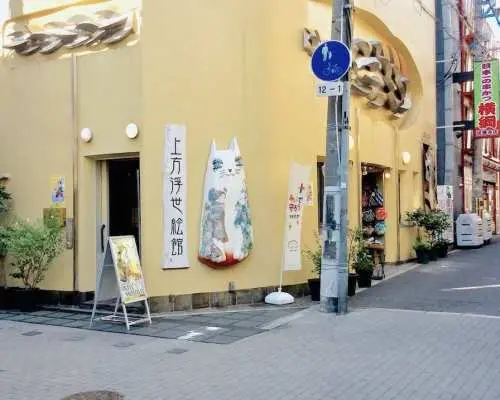 大阪上方浮世繪館街角外觀，黃色建築、招牌與特色貓咪裝飾融入街景，邀請旅客探索浮世繪藝術。