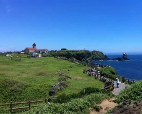 済州島「ソプチコジ」の広大な景観。緑豊かな丘陵地と海が調和し、ドラマロケ地の象徴的な建物と海岸沿いを散策する観光客。