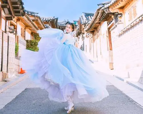 A woman joyfully twirling in an elegant light blue and purple gradient Hanbok on a picturesque traditional street in Seoul, capturing dynamic moments of the outdoor photoshoot.
