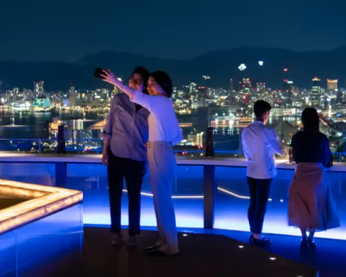 神戸屋上テラス「そらふね」で、2人の女性が壮麗な神戸の夜景と港を背景に自撮り。ロマンチックなデートの瞬間を完璧に捉えています。