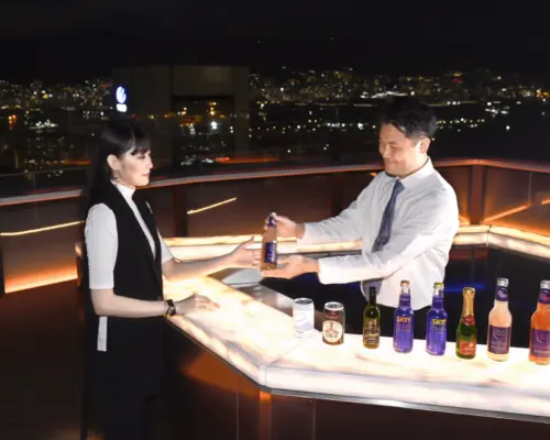 神戸屋上テラス「そらふね」で、スタッフがお客様にドリンクを提供。輝くバーカウンターと背景の神戸の夜景が、特別な高空バー体験を演出します。