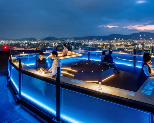 神戸「そらふね」屋上テラスの夜景。青い光で彩られた空間で、カップルや友人が110m上空から神戸市街と六甲山の輝くロマンチックな夜景を楽しんでいます。
