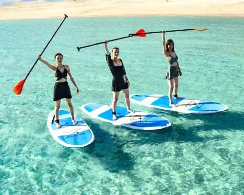 三位女性在宫古岛清澈的宫古蓝海水中体验SUP立桨冲浪，她们开心互动并摆拍，展现水上活动的乐趣与活力，背景海面波光粼粼。