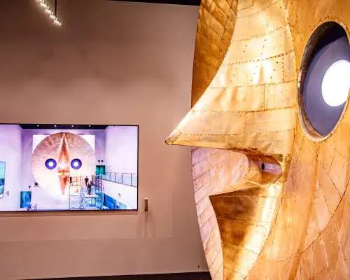 太阳之塔内部展厅，巨大的金色面孔雕塑近景，展现冈本太郎独特艺术风格，旁有屏幕播放内部结构影片。