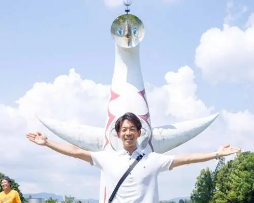 游客在大阪万博纪念公园著名的太阳之塔前兴奋留影，张开双臂拥抱旅程，体验1970年万博的历史氛围。