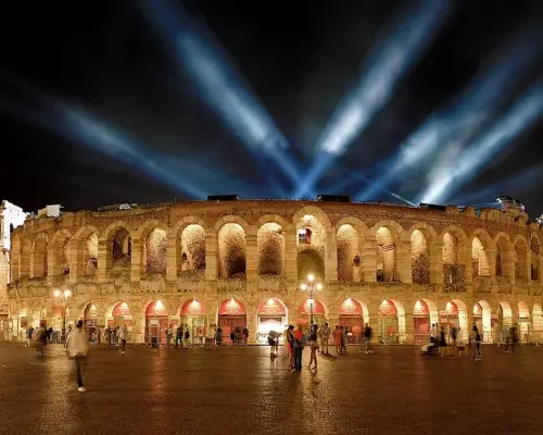 Captivating night view of the Verona Arena, illuminated by spotlights, showcasing ancient Roman ruins with visitors strolling in the piazza, offering free entry with the Verona Card.