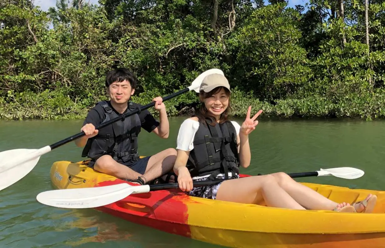 一對情侶在沖繩億首川紅樹林中划獨木舟，穿著救生衣開心地比著V字手勢，享受著平靜的水面與茂密綠色植被環繞的大自然體驗。