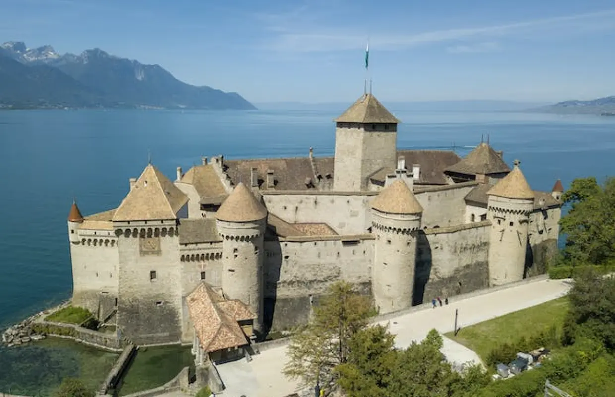 Aerial view of Chillon Castle in Montreux, Switzerland, situated on Lake Geneva, showcasing its majestic architecture under sunny skies with distant mountains and clear lake waters.