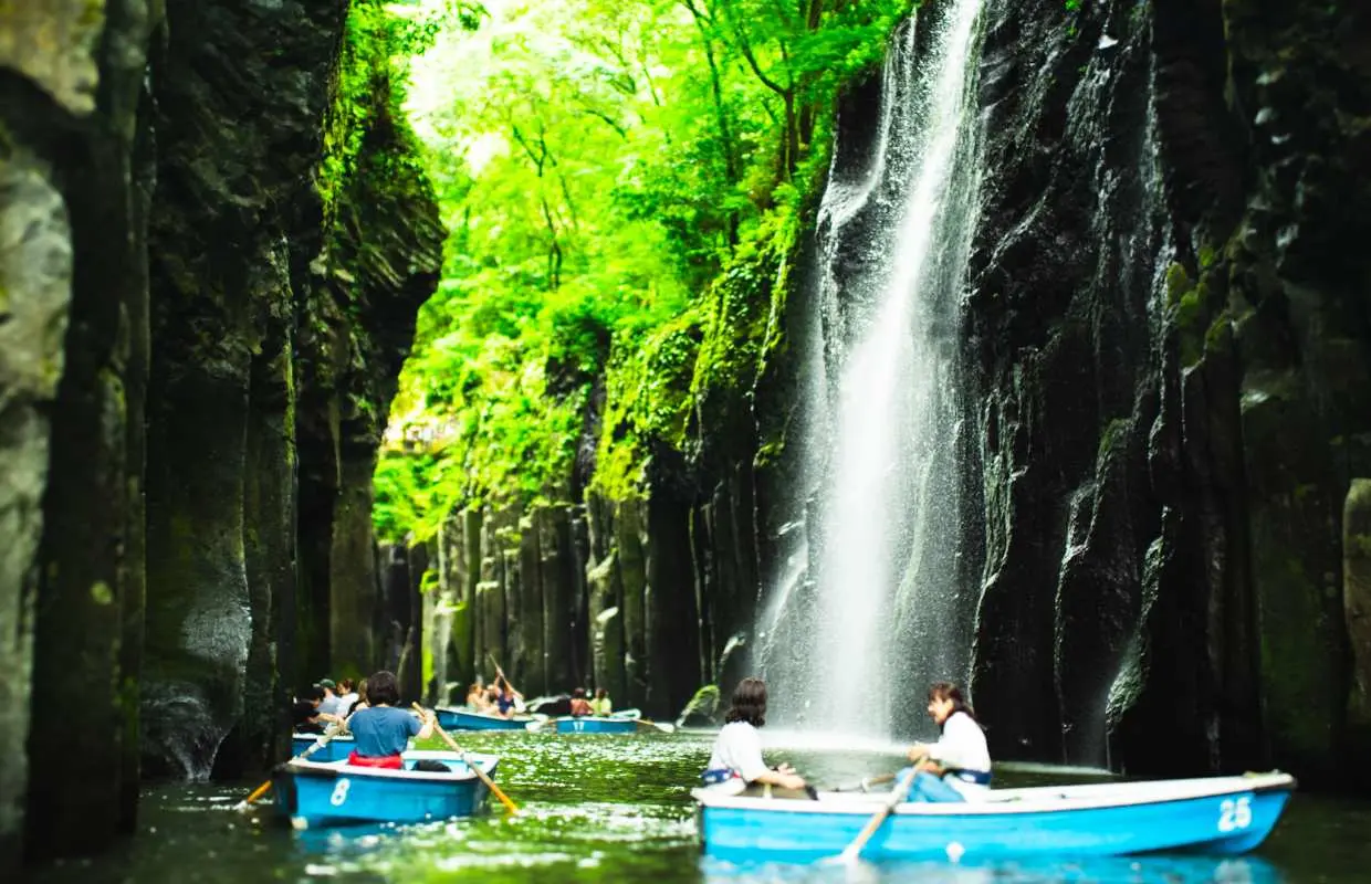 高千穂峡でのボート体験、観光客が真名井の滝に近づき、水しぶきを感じながら、両側の緑豊かな崖に囲まれた自然の絶景を体験。