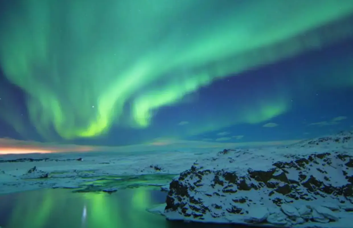 冰島雷克雅維克夜空下，綠色極光舞動在雪地與結冰湖泊上，倒映出夢幻光影，呈現極光船之旅的壯麗景色。