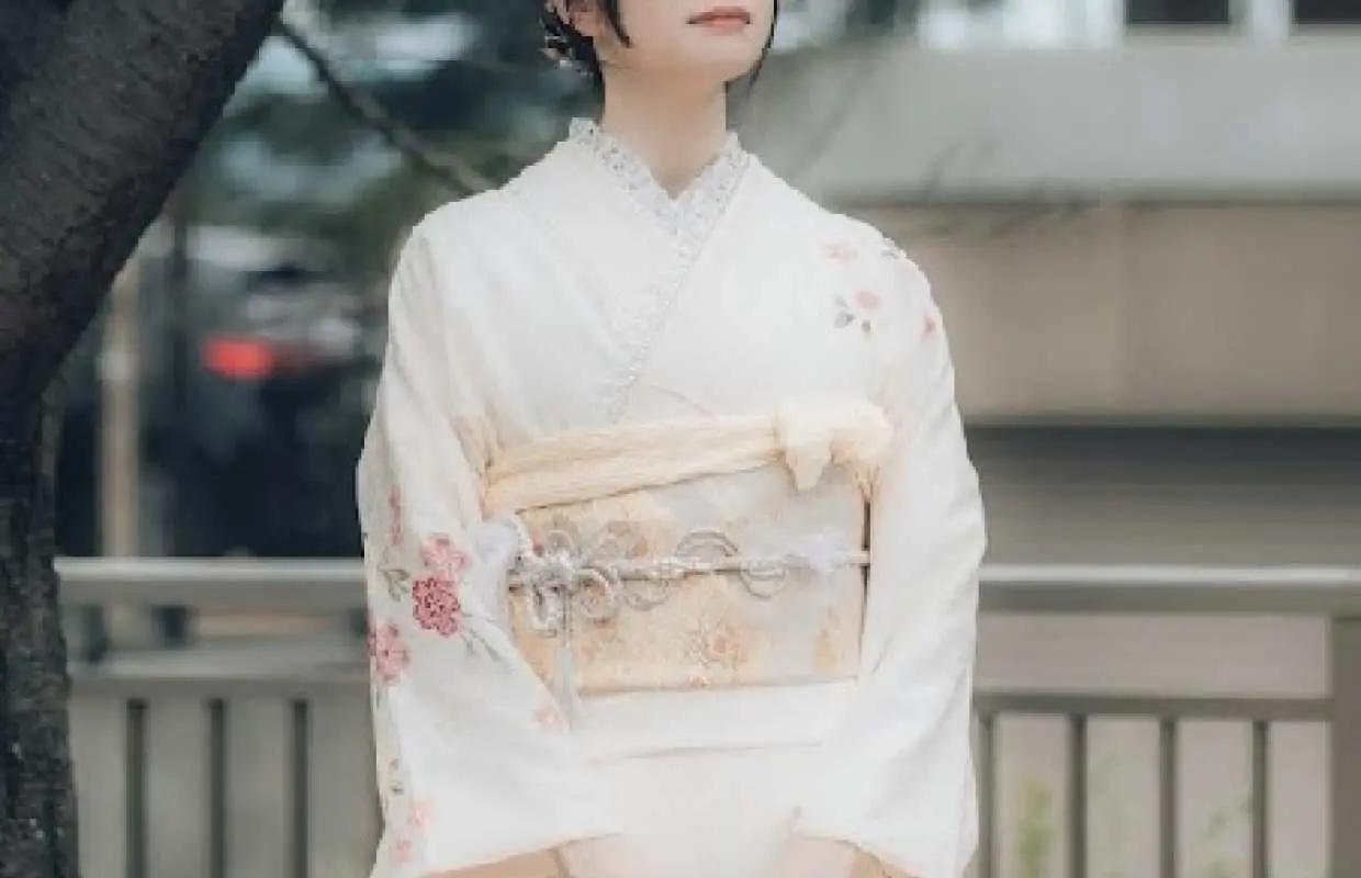 日本京都和服體驗，女性身著素雅白色和服，袖口與裙擺飾有精緻花卉，在綠樹下優雅佇立，展現日式美學。