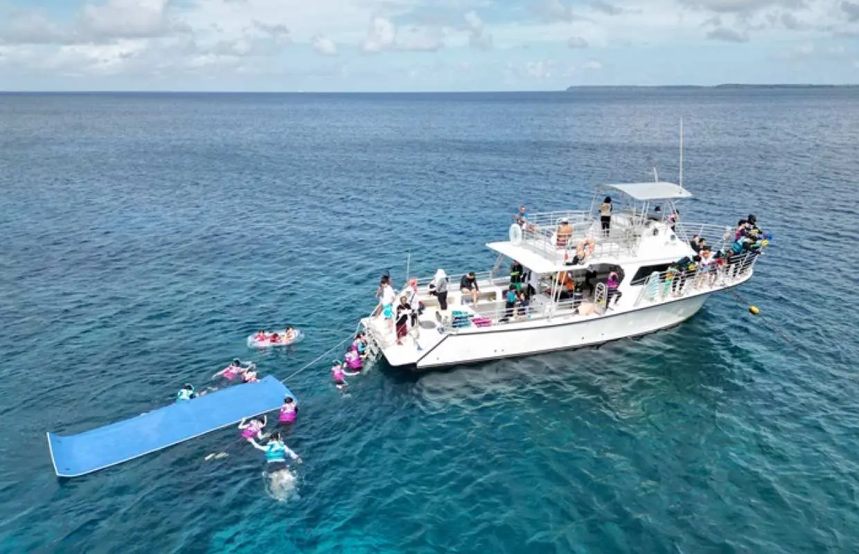 Aerial view of the lively Guam Princess Dolphin Cruise yacht, with guests snorkeling, enjoying an inflatable slide, and water activities in the clear Pacific ocean.
