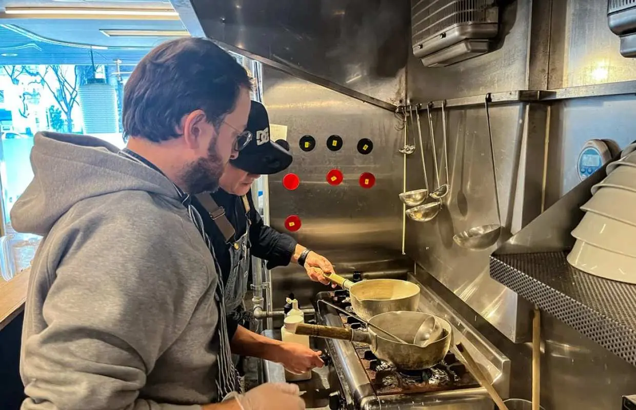 A student learning to cook authentic chicken broth ramen with professional ramen chef Shu in a stainless steel kitchen during an Osaka ramen cooking class.