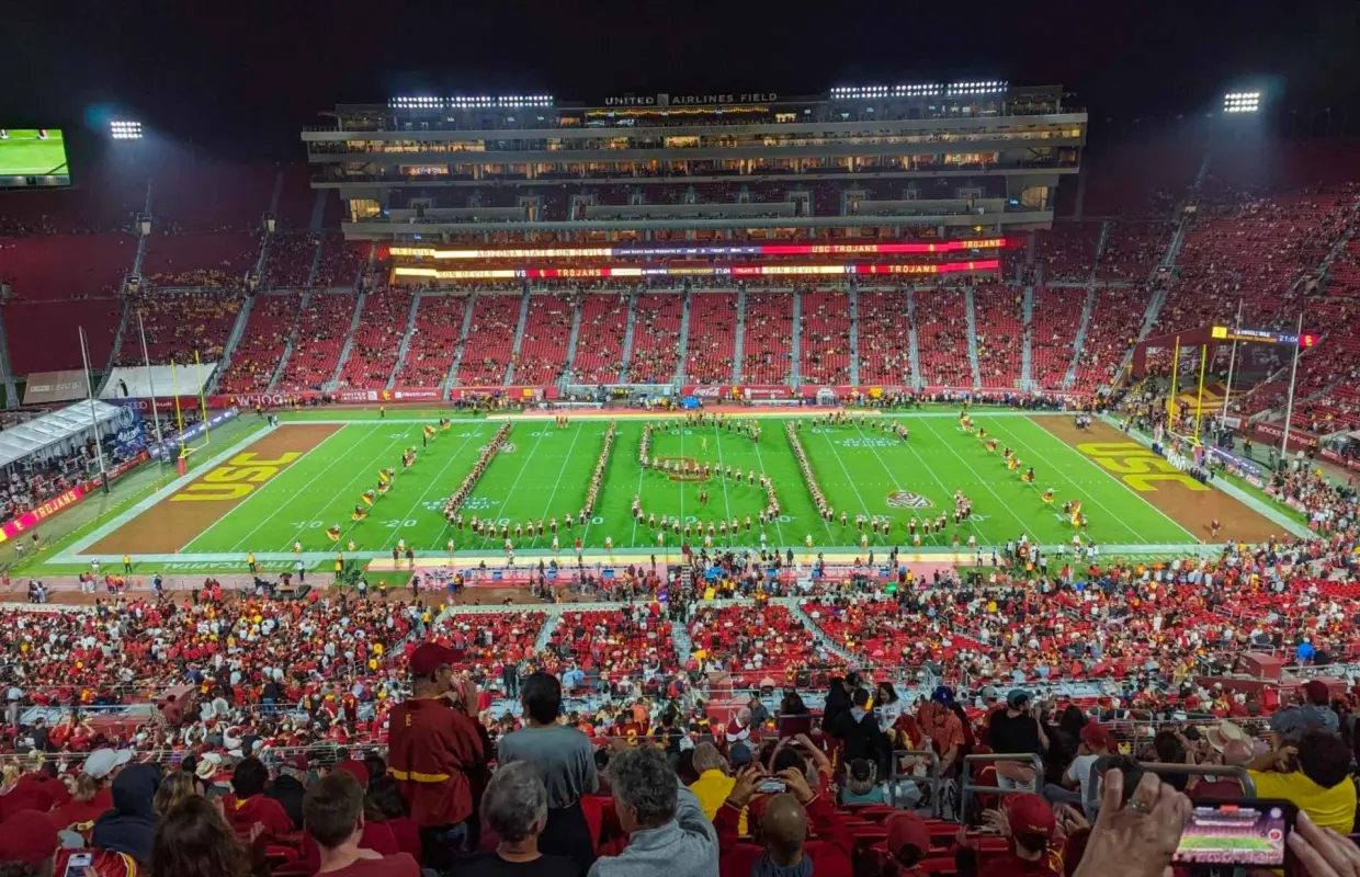 Toàn cảnh trận bóng bầu dục đêm của đội USC Trojans tại Sân vận động Tưởng niệm Los Angeles, với ban nhạc diễu hành tạo hình 'USC' trên sân và hàng ngàn người hâm mộ trên khán đài tận hưởng không khí sôi động.