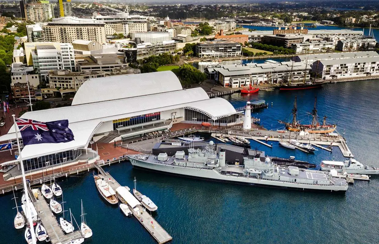 Aerial view of the Australian National Maritime Museum in Sydney's Darling Harbour, featuring the main building, a docked destroyer, sailboats, and the city skyline, highlighting Australia's maritime history.