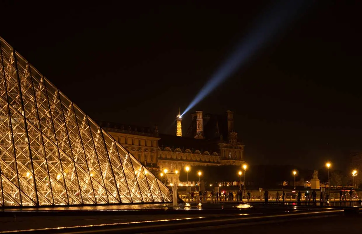 The magnificent Louvre Pyramid illuminated at night, with the iconic Eiffel Tower's light beam shining across the Parisian skyline, a highlight of the night tour.