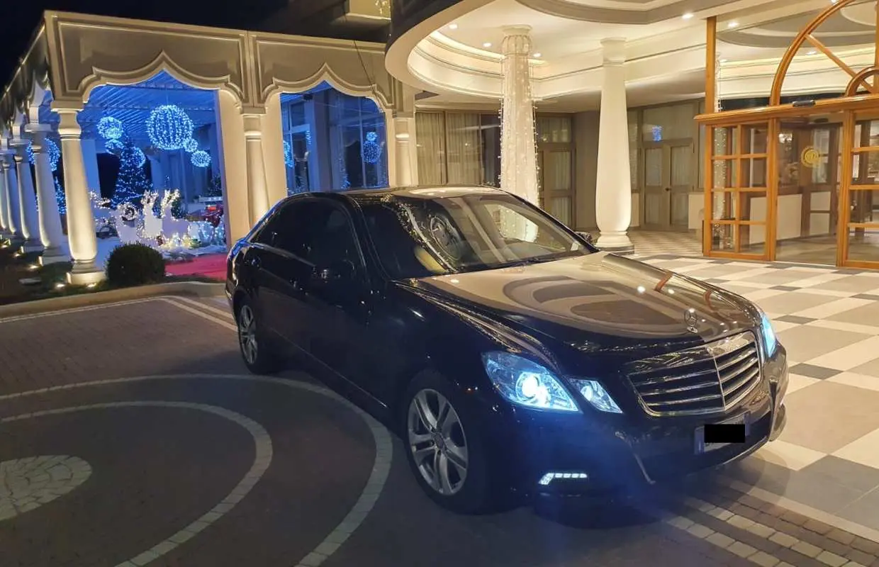 一辆黑色奔驰豪华轿车停在埃因霍温酒店入口处，夜间灯光璀璨，提供便捷的机场私人接送服务。