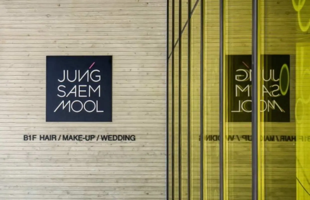 Exterior view of JUNG SAEM MOOL Inspiration West branch in Seoul, featuring its distinctive logo on a wooden wall and reflecting glass, highlighting hair, makeup, and wedding services near Apgujeong Rodeo Station.