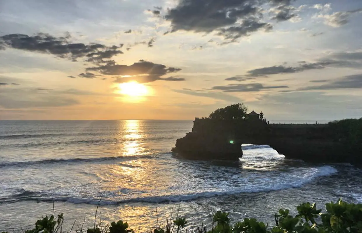 バリ島タナロット寺院の夕日。夕焼け空と海を背景に、波が打ち寄せる岩礁に立つ神秘的な寺院の絶景。