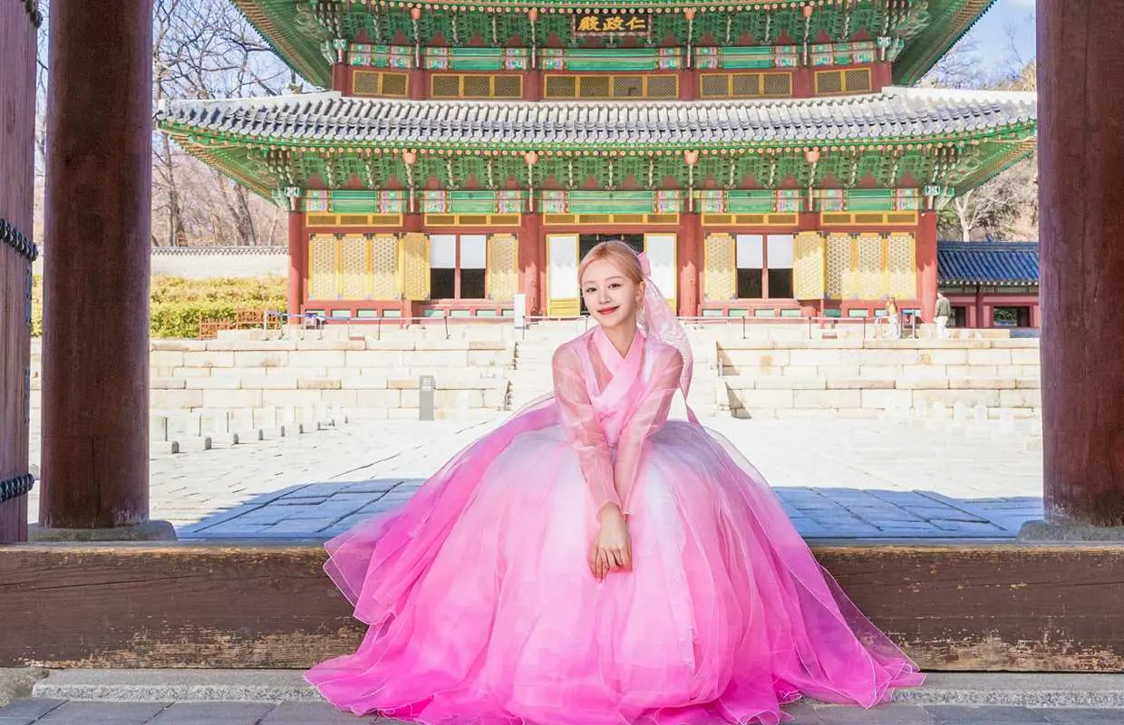 A woman in a beautiful pink gradient Hanbok elegantly seated in front of Seoul's majestic Changdeokgung Palace, enjoying a traditional Hanbok photoshoot experience.
