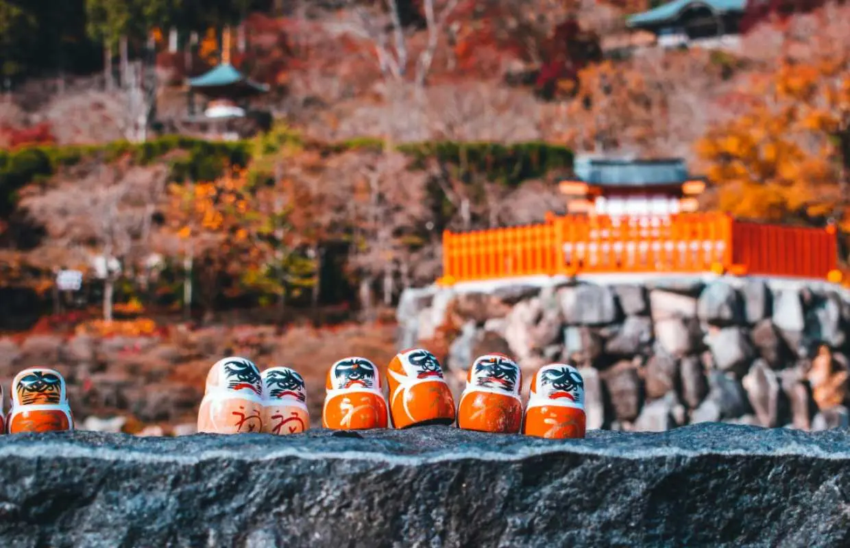 大阪胜尾寺秋日景色，达摩不倒翁整齐排列于石墙上，背景是枫红与古老寺院，充满日式风情。
