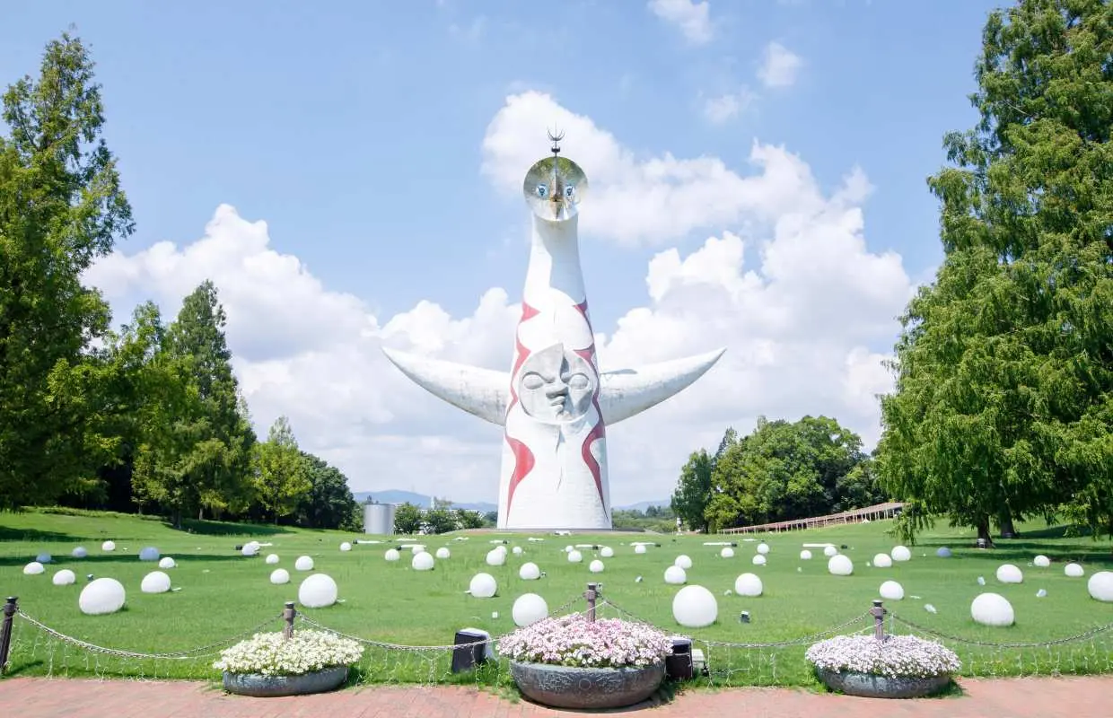 大阪万博纪念公园中，冈本太郎著名的太阳之塔在蓝天绿地下雄伟矗立，周围点缀着白色灯球与茂盛树木。