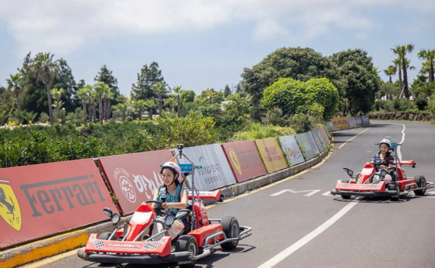 Two racers in red go-karts navigate the thrilling Wind 1947 extreme track in Jeju, passing a Ferrari-branded barrier amidst lush green trees, enhancing the racing experience with an adrenaline rush.