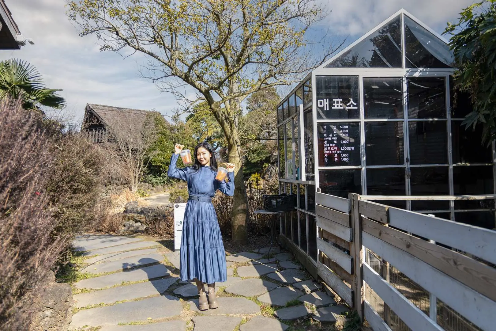 A woman strolling along a stone path at Jeju Aewol Sheep Ranch, with a modern glass building and natural trees in the background, showcasing the ranch's blended environment.