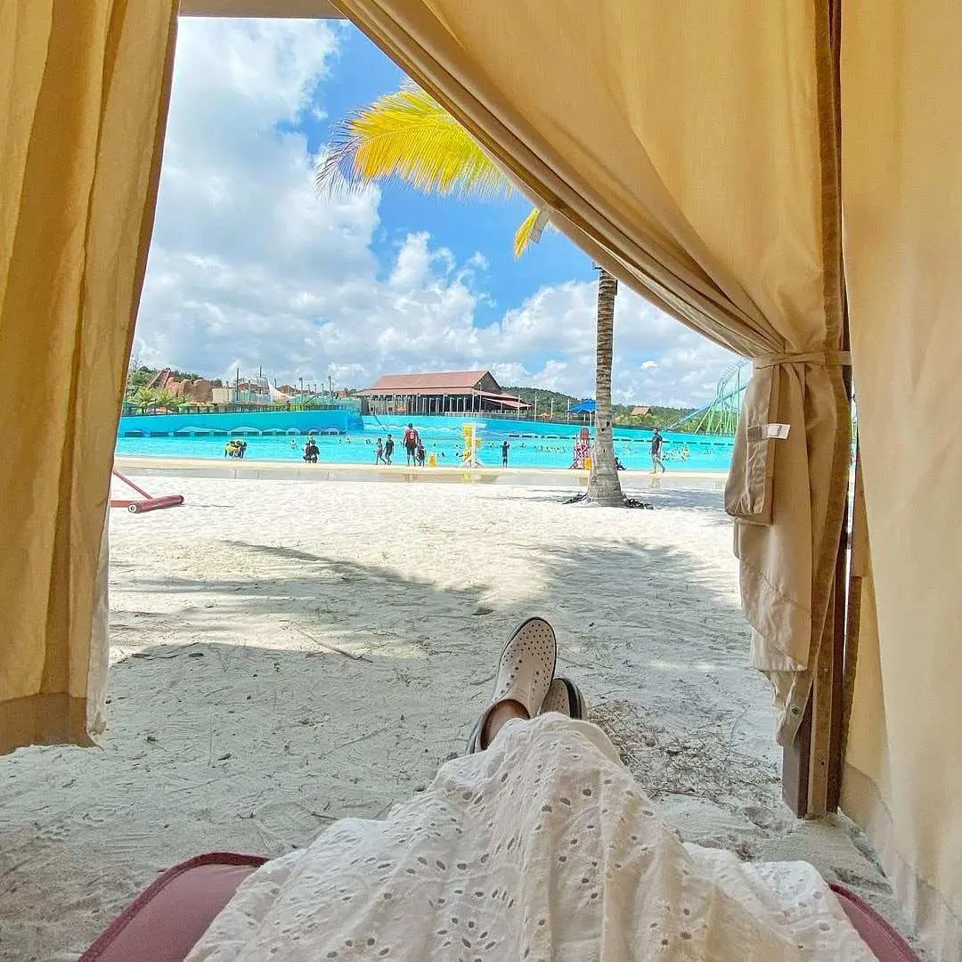 A relaxing view from inside a private cabana at Desaru Coast Adventure Waterpark, looking out towards the bustling wave pool, sandy beach, and palm trees under a bright blue sky.