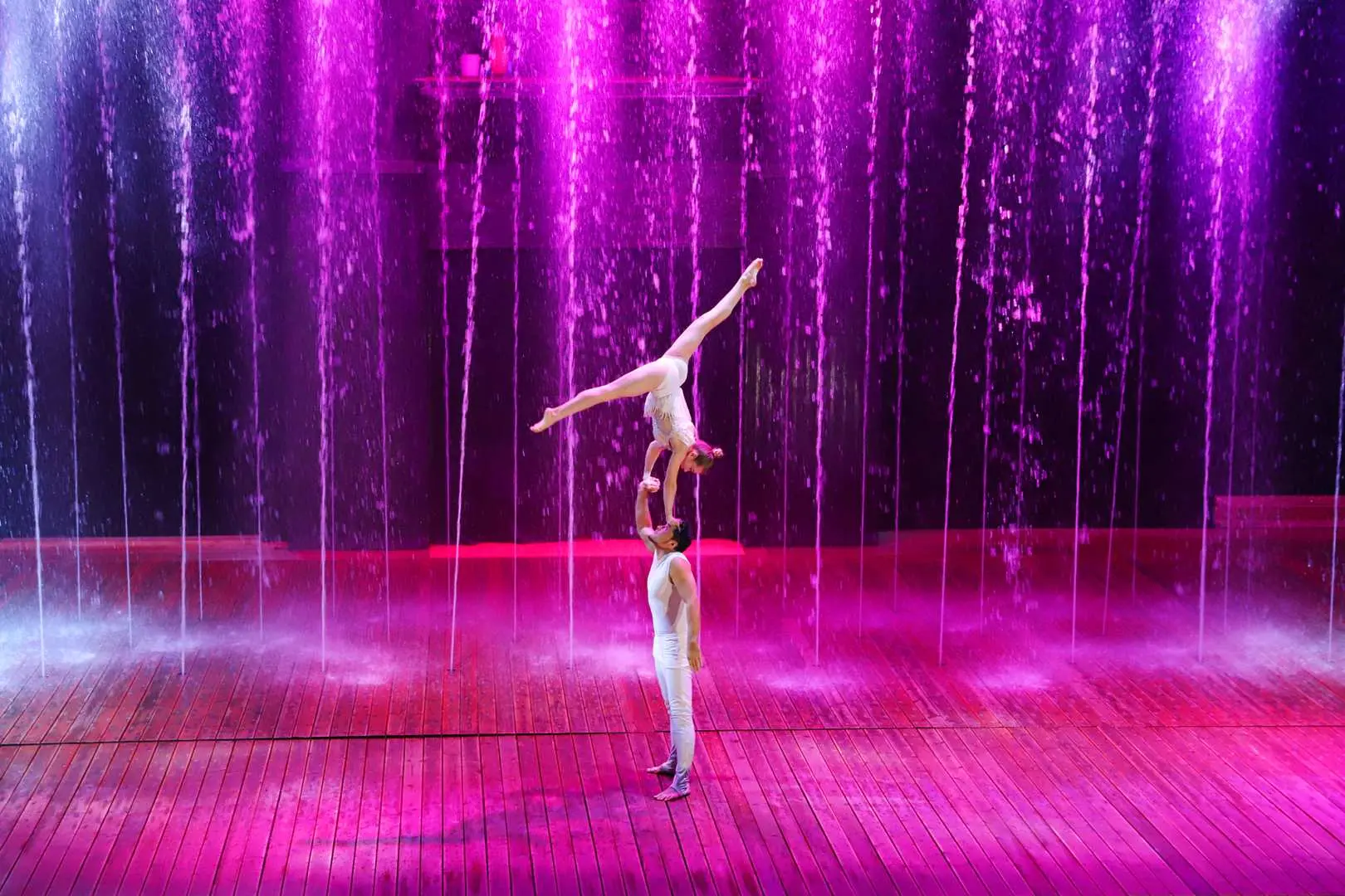 Jeju Island Sky Water Show, two acrobats perform a high-difficulty duo balancing act in front of a purple water curtain, with one person hand-standing on the other, viewed from the audience's perspective.