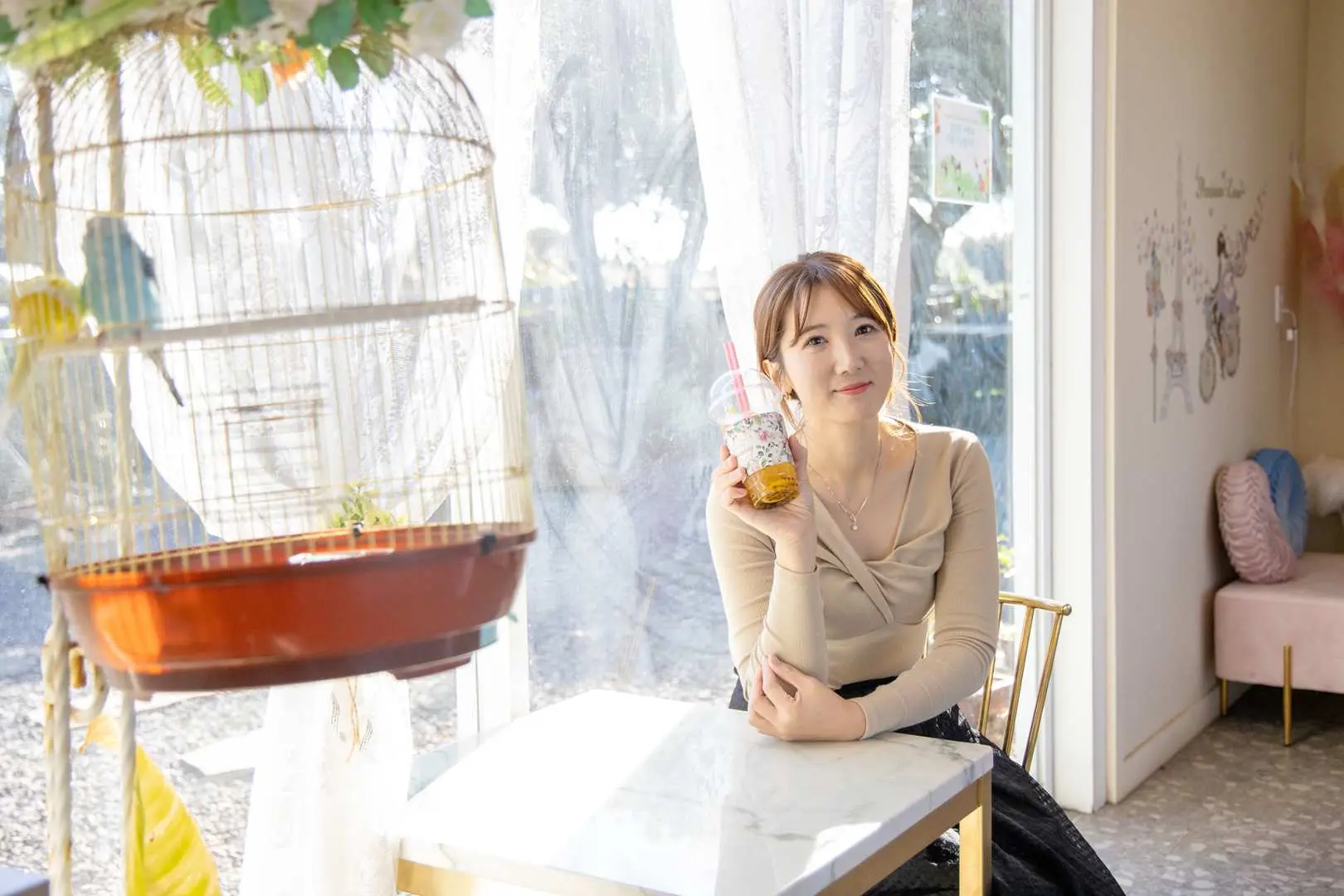 제주도 플라워 동물원의 밝은 카페 창가에서 한 여성 방문객이 음료를 들고 미소 짓고 있으며, 옆에는 귀여운 새장이 놓여 있습니다.