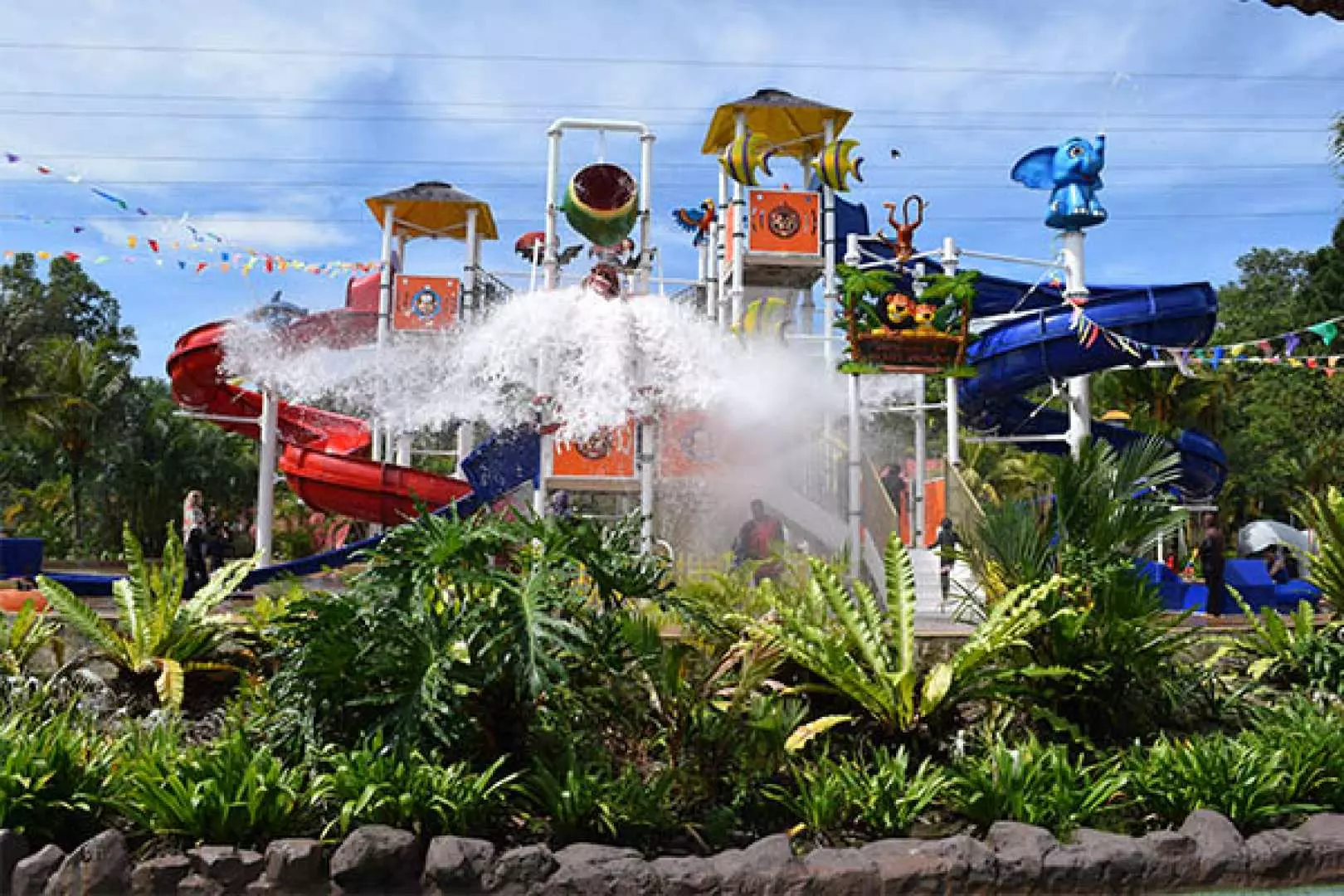 A large water playground at Bukit Merah Laketown Waterpark in Perak, Malaysia, featuring a huge tipping bucket and various slides, surrounded by lush tropical plants, offering a refreshing experience.