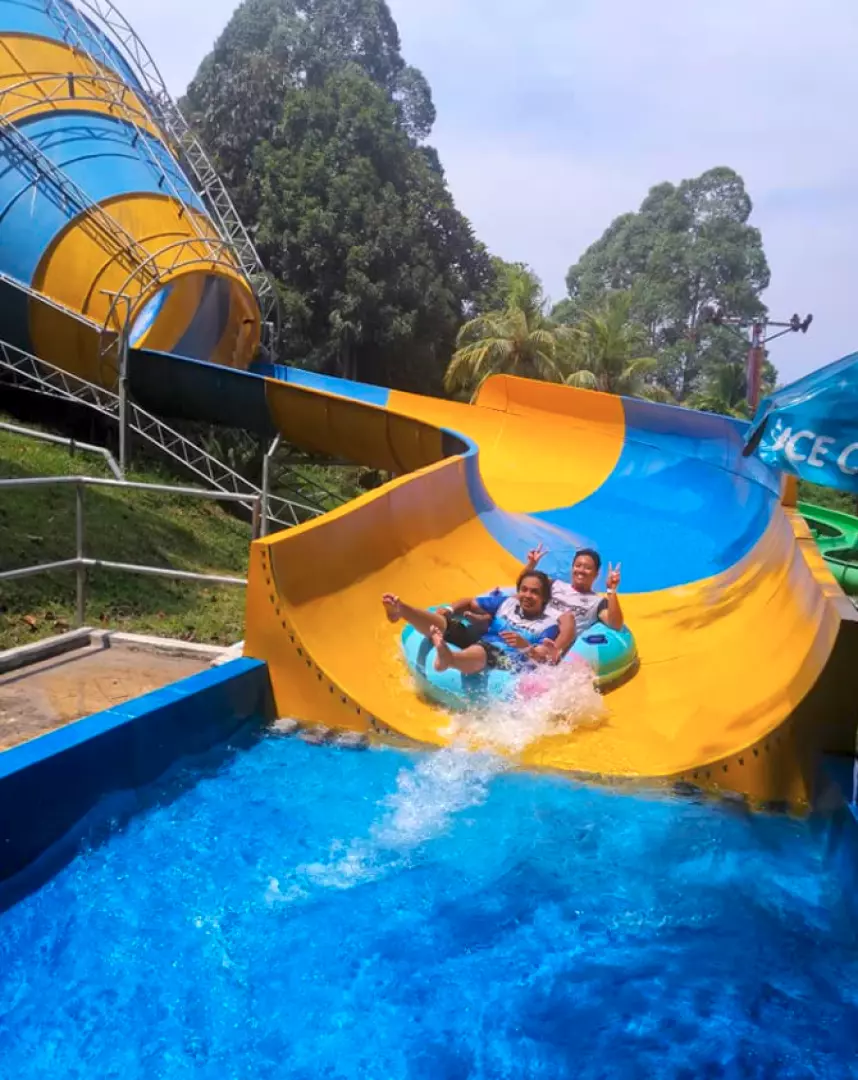 Two visitors on an inflatable tube splash out of a blue and yellow spiral water slide at Bukit Merah Laketown Waterpark in Perak, Malaysia, enjoying a thrilling ride together.