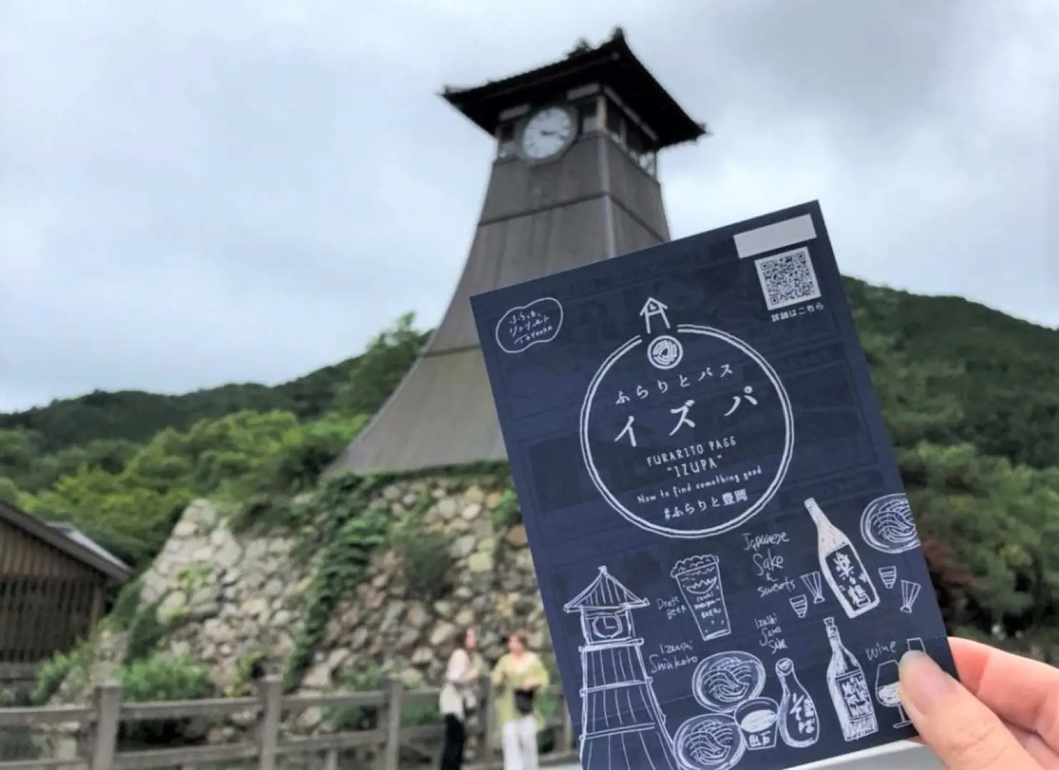出石城下町の象徴である辰鼓楼の時計台と緑豊かな丘を背景に、「イズパ」通行券パンフレットを持つ手。兵庫での便利な観光の魅力を強調しています。