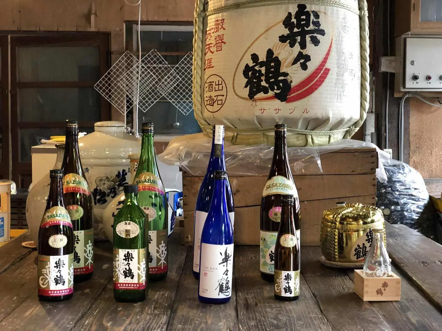 様々なサイズとラベルの日本酒ボトルが、大きな伝統的な酒樽を囲むように並んでいます。出石城下町「イズパ」通行券で楽しめる本格的な日本酒試飲体験を紹介。