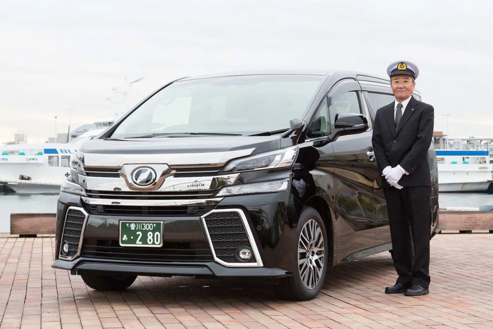 A professional driver in uniform stands smiling by a luxurious black Alphard/Vellfire minivan, offering private chartered tours for sightseeing in Shikoku, Kagawa.