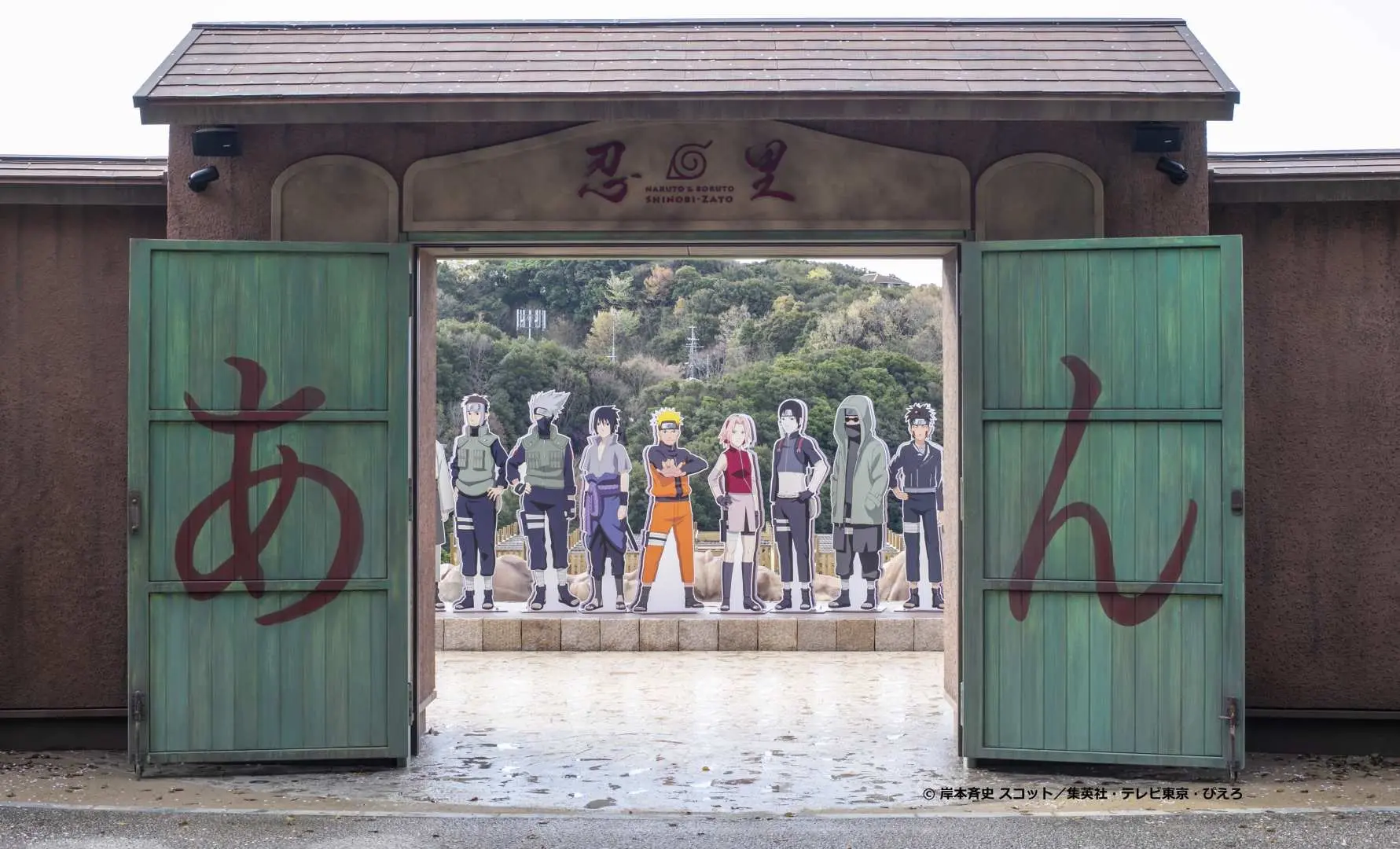 The entrance to Awaji Island's Nijigen no Mori 'NARUTO & BORUTO Shinobi-Zato' theme park, featuring green wooden doors with 'Shinobi-Zato' written on them, and Naruto character standees visible inside.