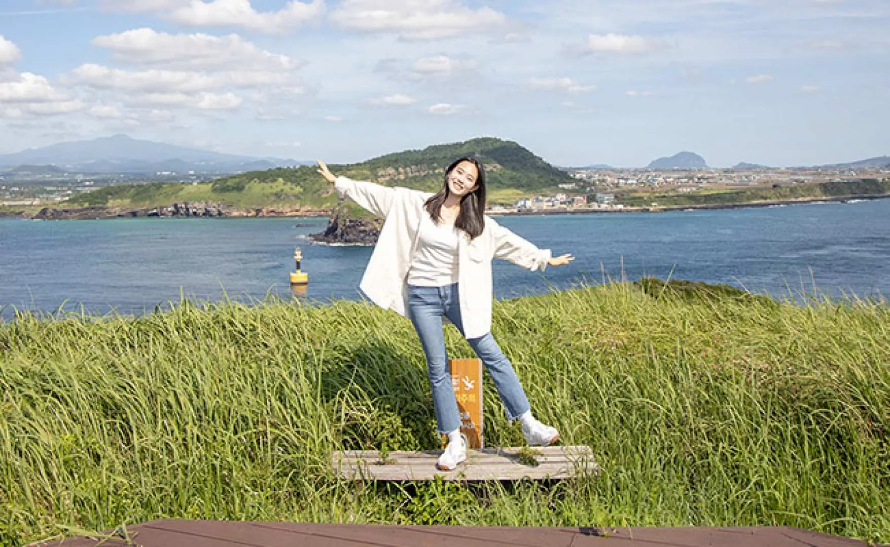 제주 차귀도 해안가 풀밭에서 젊은 여성이 환하게 웃으며 팔을 벌리고 서 있고, 뒤로는 푸른 바다, 작은 등대, 멀리 마을이 보입니다.