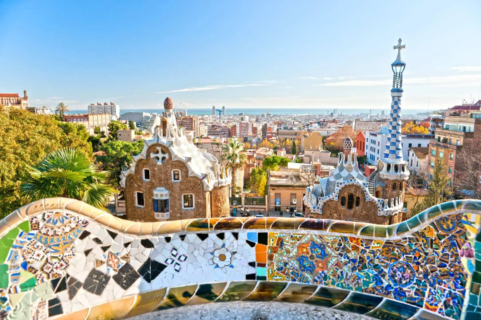 Majestic daytime panoramic view from Park Güell's main plaza in Barcelona, featuring the colorful mosaic bench and Gaudí's fairytale gatehouses, with visitors enjoying the city and sea views.
