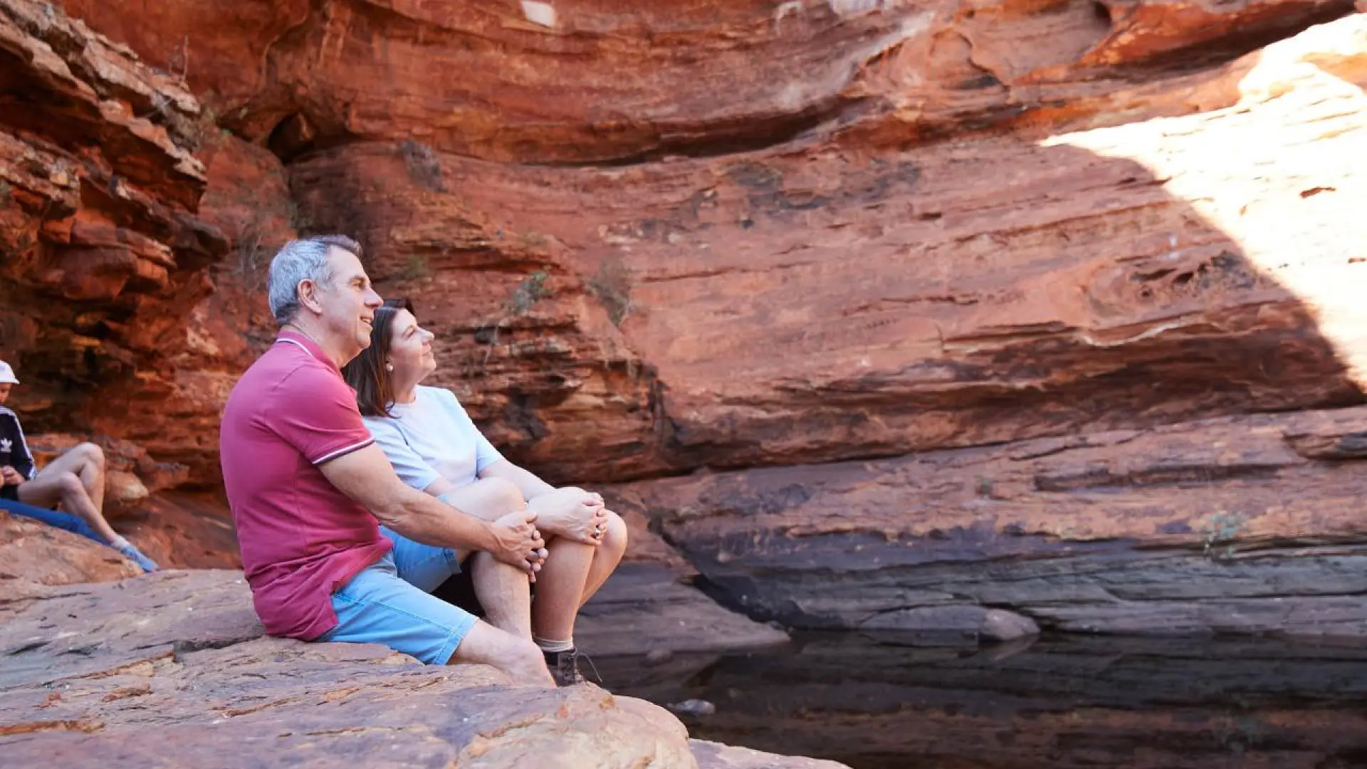 A couple sits by a natural water pool in Kings Canyon's Garden of Eden, surrounded by magnificent red sandstone cliffs, enjoying the tranquility and beauty of this outback oasis.