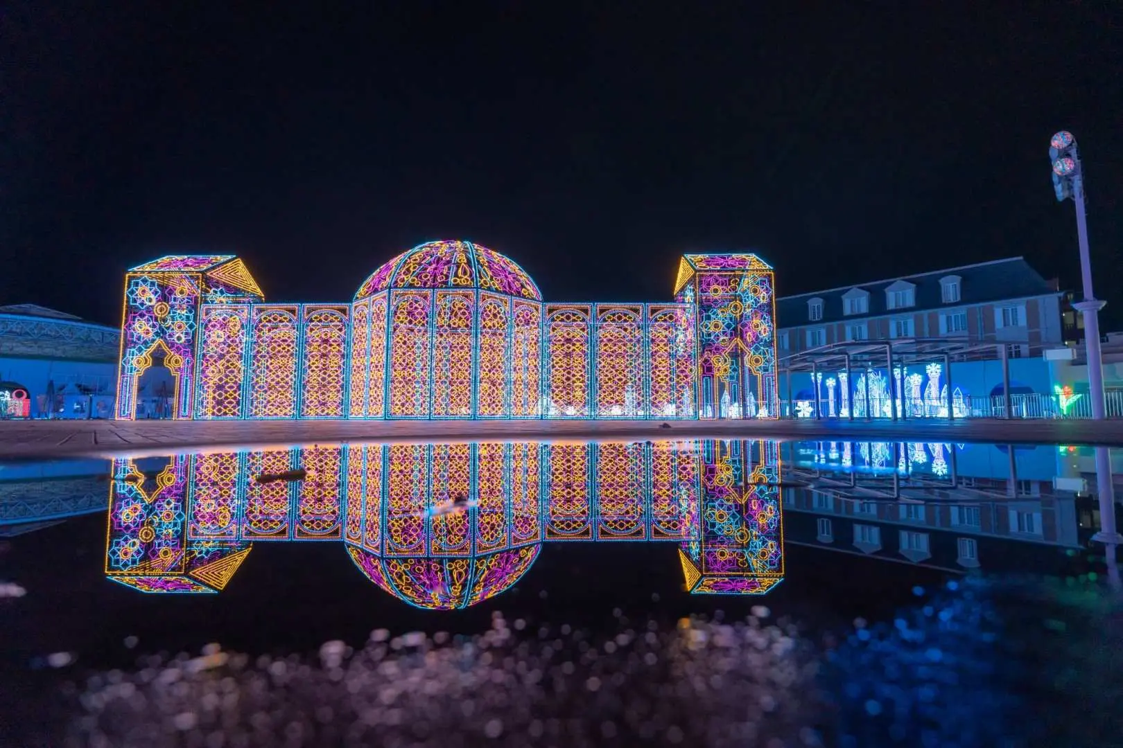 神戸イルミナージュ「夢の庭園」のテーマライトアップを側面から見た景色。華やかな色彩の光の建物が夜空に輝き、静かな水面に鮮やかに反射しています。