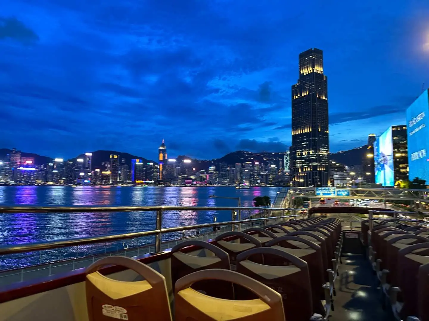 オープントップバスの2階から香港ビクトリアハーバーの夕暮れから夜にかけての景色を眺めます。都市のスカイラインが深い青空の下で輝き、夜間観光を楽しむ準備ができています。