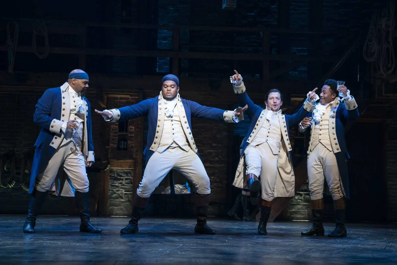 Live performance shot of the Broadway musical 'Hamilton' featuring four male actors in American Revolutionary War era costumes, energetically singing and dancing on stage.
