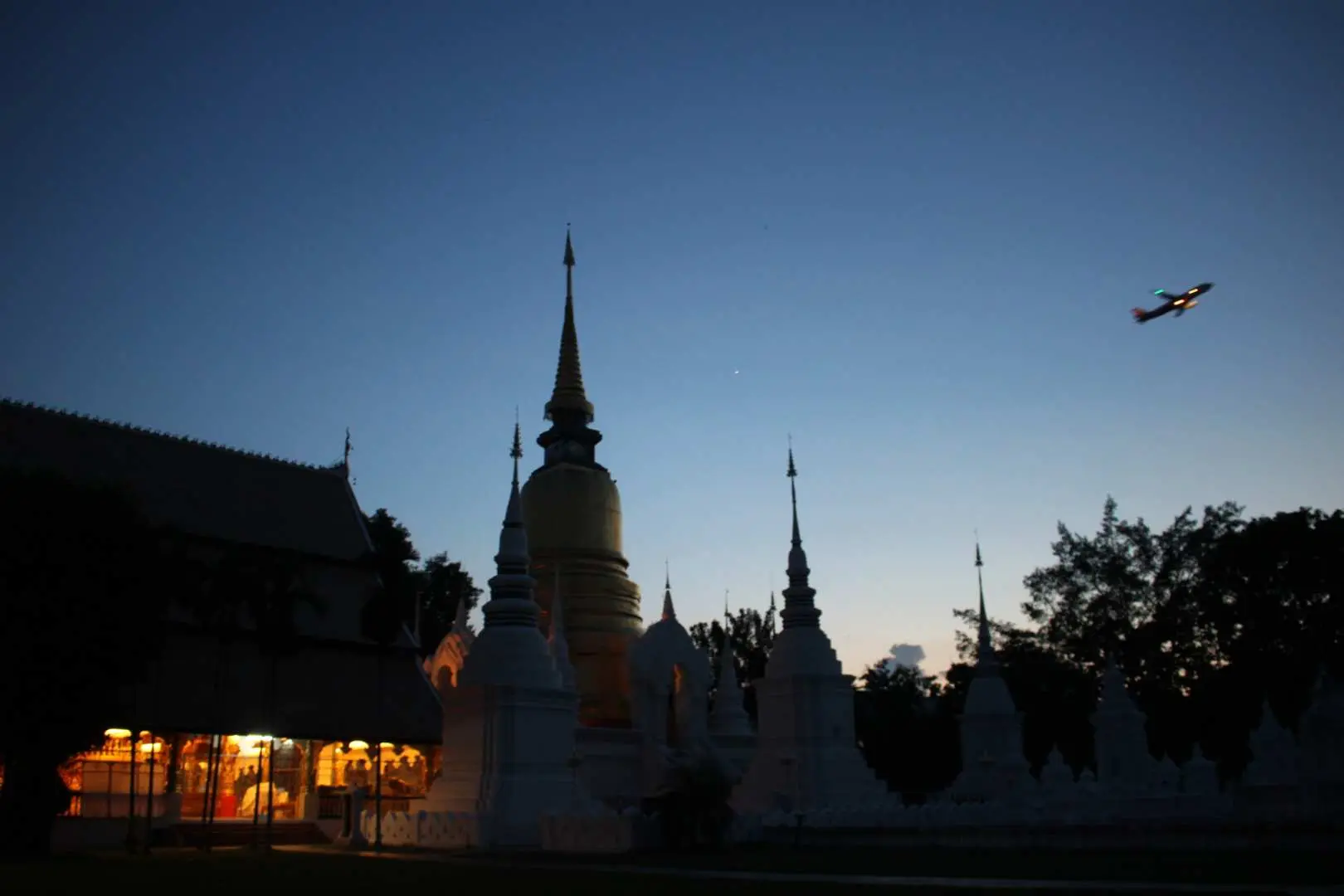 清邁蘇安杜克寺在日落暮色中，金頂佛塔與白色舍利塔群靜謐莊嚴，一架飛機劃過天際，為嘟嘟車夜遊增添獨特風景。
