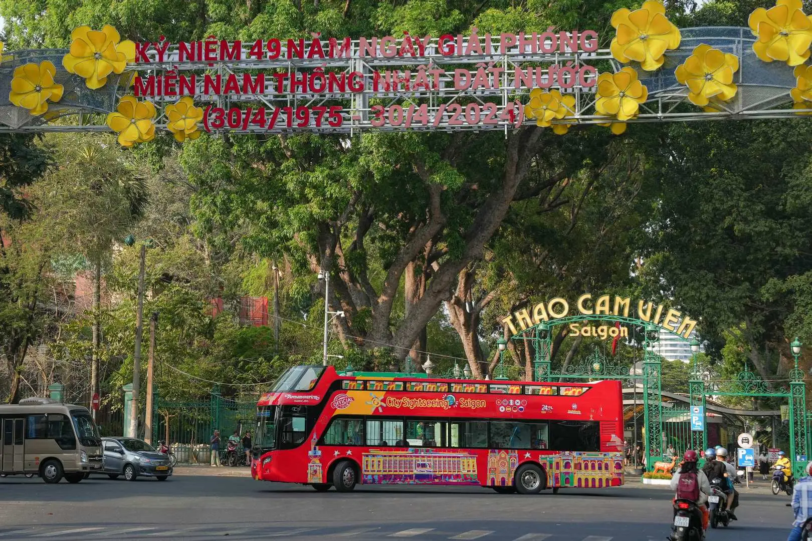 The Ho Chi Minh City hop-on hop-off double-decker sightseeing bus passes near the entrance of Saigon Zoo and Botanical Garden, with a decorative archway above, showcasing city attractions along the Saigon tour route.