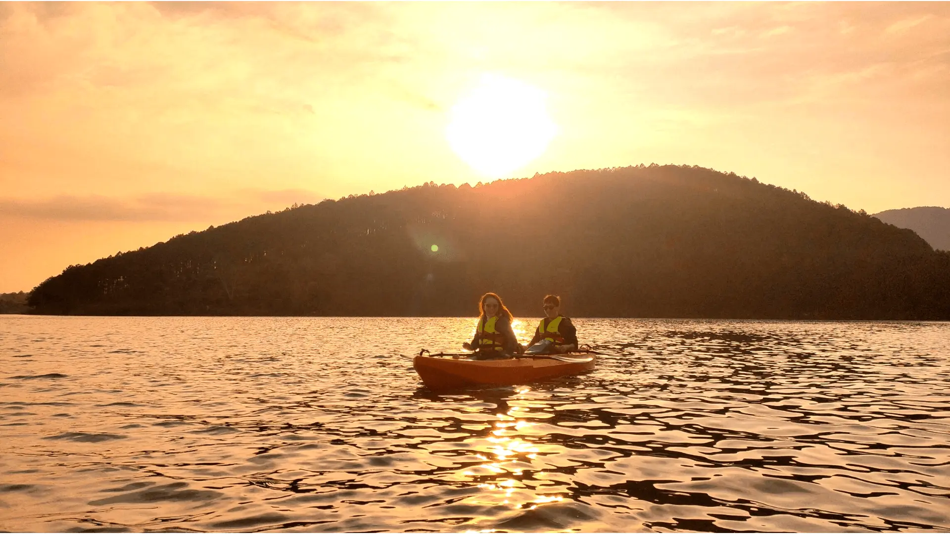 Hai người chèo thuyền kayak về phía núi non dưới ánh hoàng hôn trên hồ Tuyền Lâm, mặt nước lấp lánh tạo nên không khí lãng mạn và yên bình.