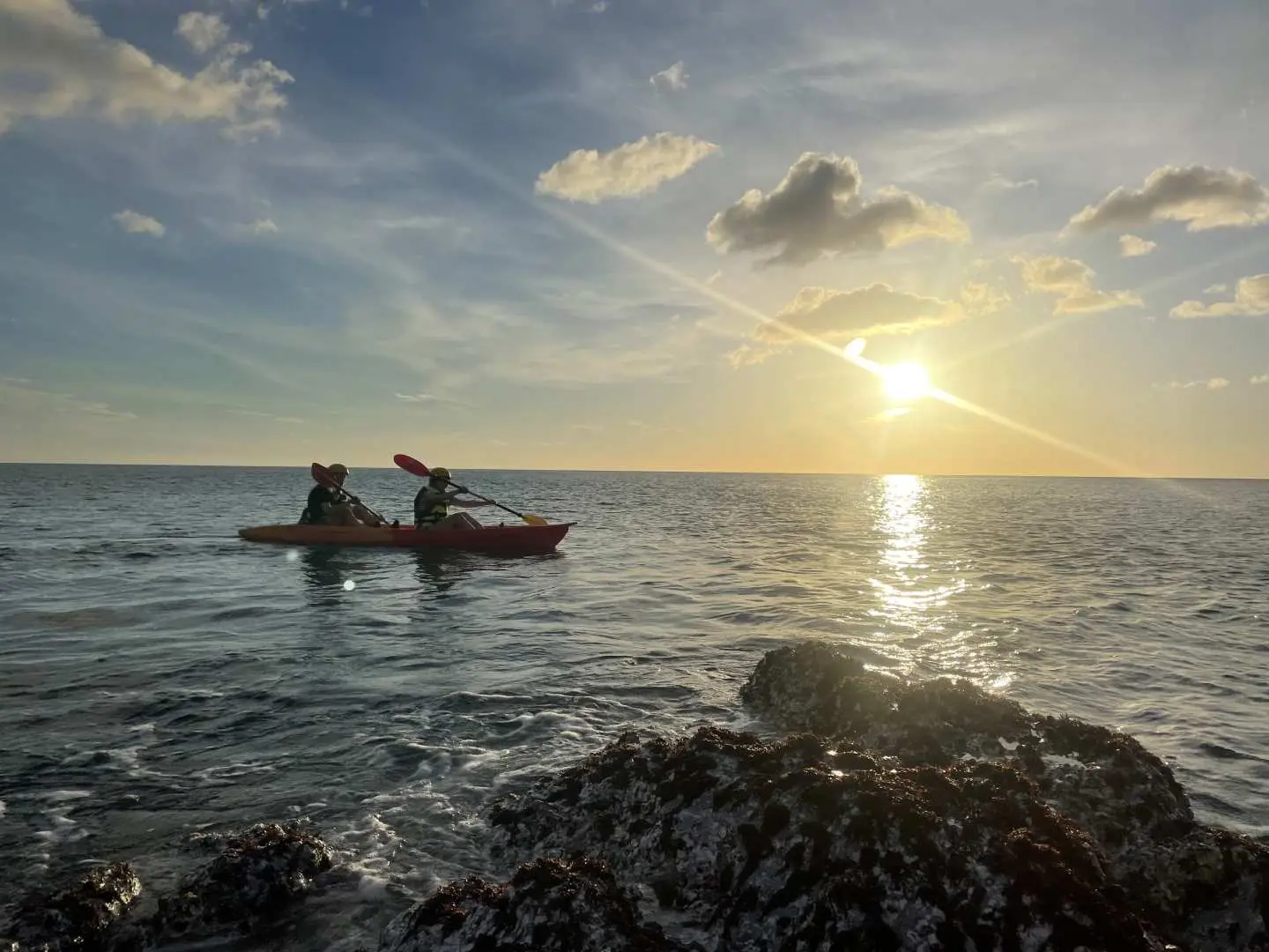 Two participants kayaking on the sunny Pingtung Alangyi sea, with sunlight reflecting on the water and rugged rock formations in the foreground, showcasing a morning or daytime marine adventure.