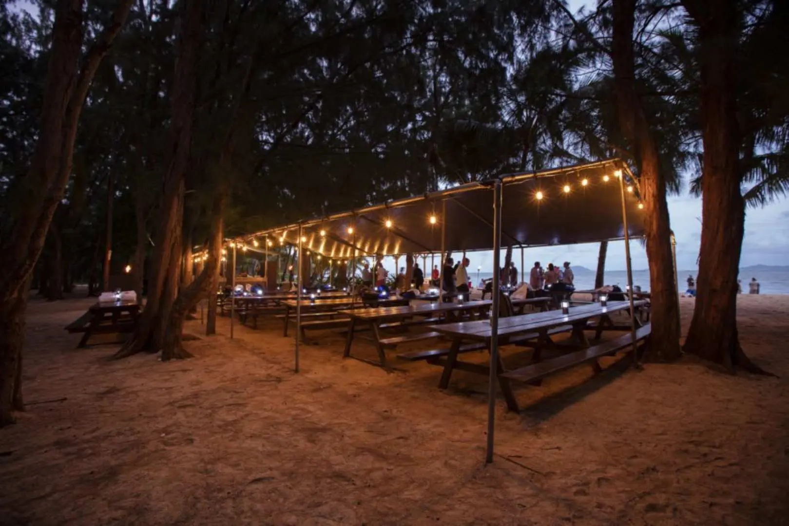 Nighttime outdoor dining area at Kualoa Secret Island Beach, Oahu, Hawaii, with festive string lights illuminating picnic tables under trees, offering a tranquil gathering experience away from crowds.